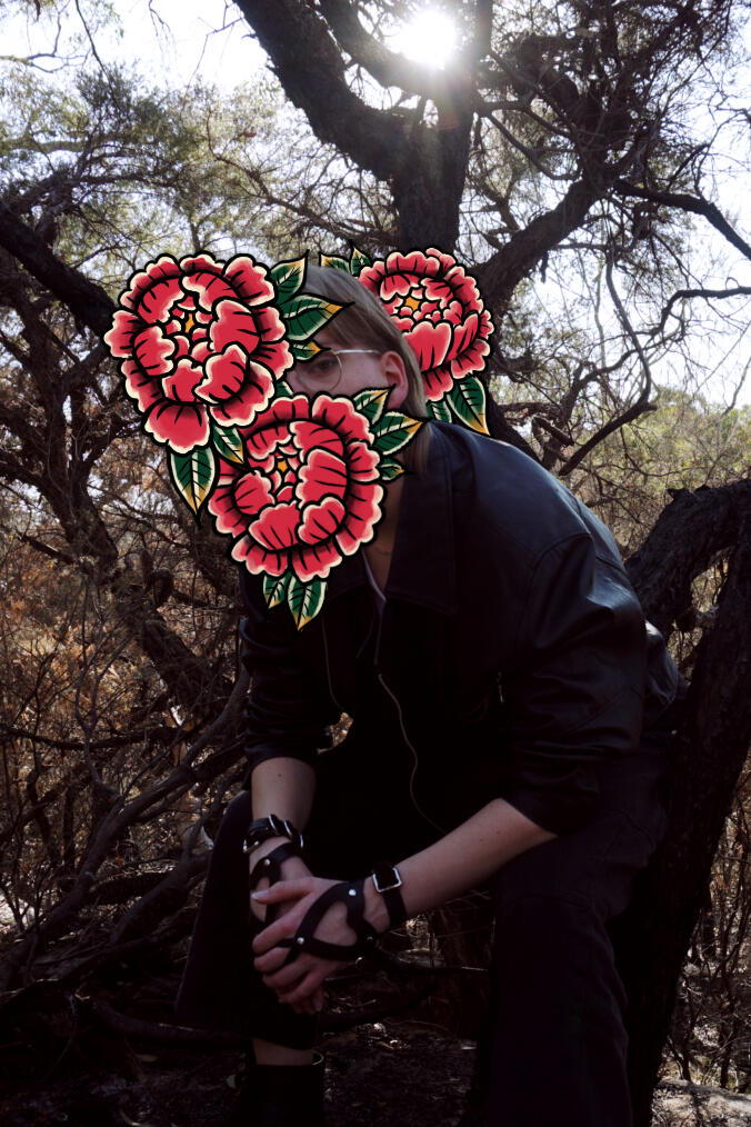 Portrait of Odile York. She is a pale skinned woman with a blonde mullet sitting on a burnt tree branch. She is wearing a leather jacket and her face is covered in illustrated flowers.